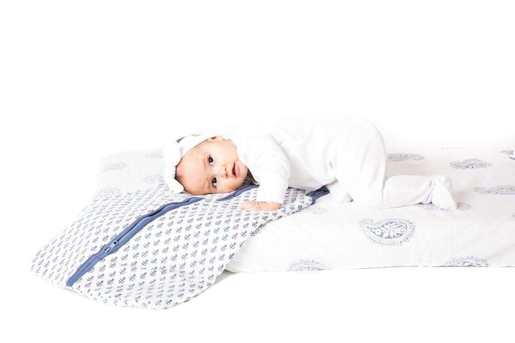 Baby lying on white and blue patterned FORT Wearable Baby Sleep Bag with zipper, dressed in white outfit and headband